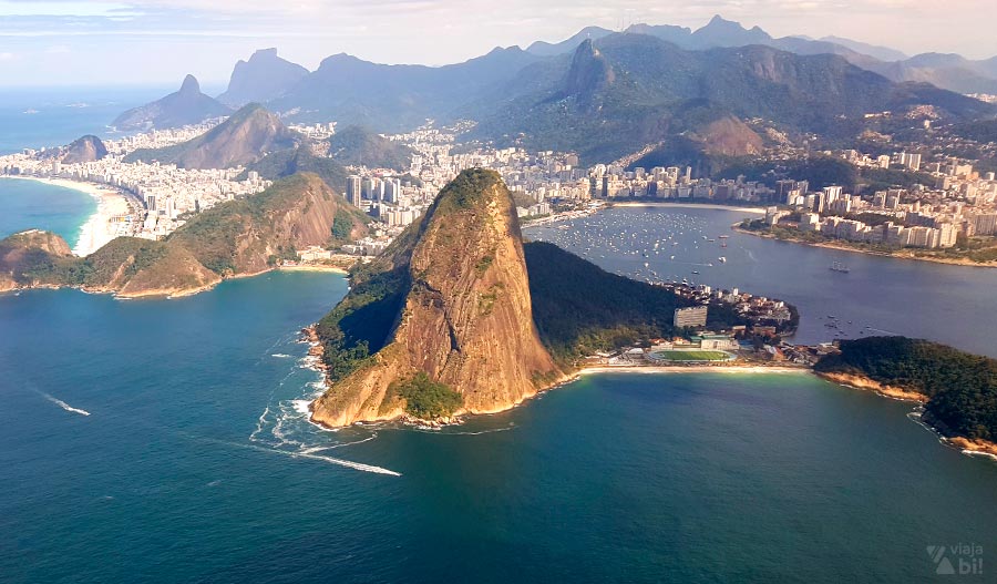 Rio de Janeiro visto do alto, com o Pão de Açúcar à frente e toda formação da cidade, entre os morros, ao fundo. É possível ver o Corcovado com o Cristo Redentor, as praias e a Pedra da Gávea.