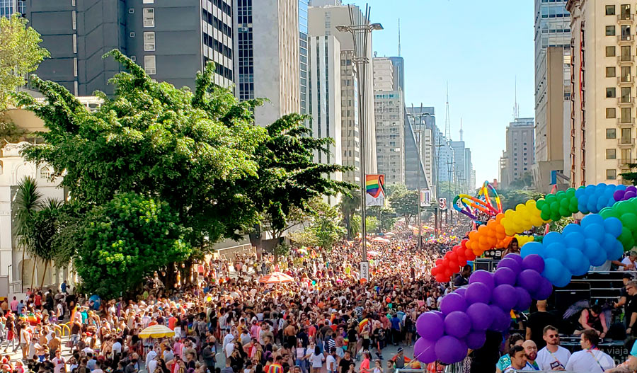 Multidão vista de cima de trio da Parada LGBTQIA+ de SP. É possível ver arcos de bexigas coloridas no trio da frente e, ao fundo, a Av. Paulista com gente a se perder de vista e o prédio da FIESP.