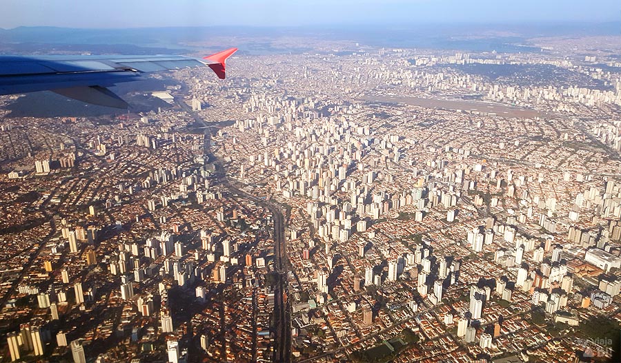 Vista de São Paulo vista de cima, da janela de um avião. Pode-se ver um pedaço da asa do avião e os diversos prédios pequenos lá embaixo, cortados por uma faixa mais escura, um dos rios da cidade.