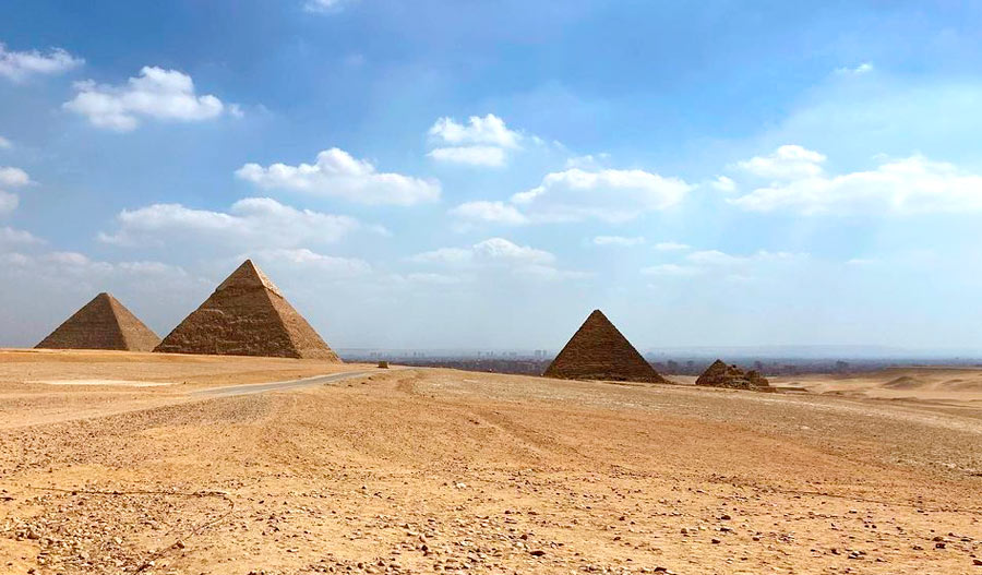 Pirâmides do Egito vistas de longe, com terreno desértico se estendendo desde onde a foto foi tirada até a atração. O céu está azul com algumas nuvens brancas.