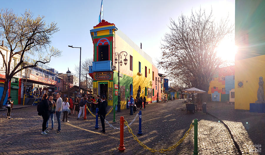 Prédio colorido característico da região de El Caminito, em Buenos Aires, colorido em diversas cores primárias. Na rua de pedestres, pessoas passam. Por trás de uma árvore, se vê o brilho do sol de fim de tarde passando.