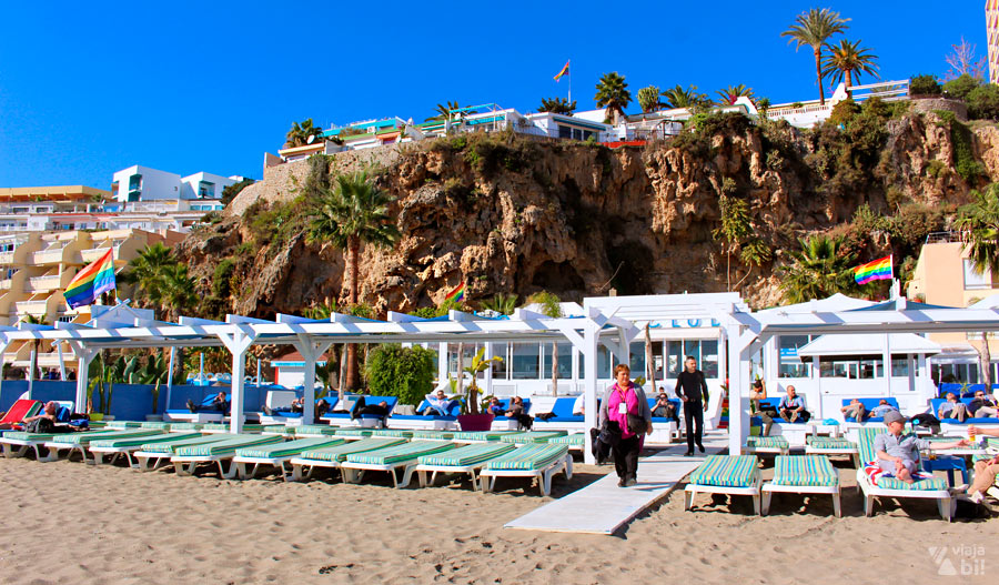 Beach club com espreguiçadeiras na areia, estrutura em madeira branca, passarelas e bandeiras do orgulho em frente a um paredão de pedra com construções no topo.