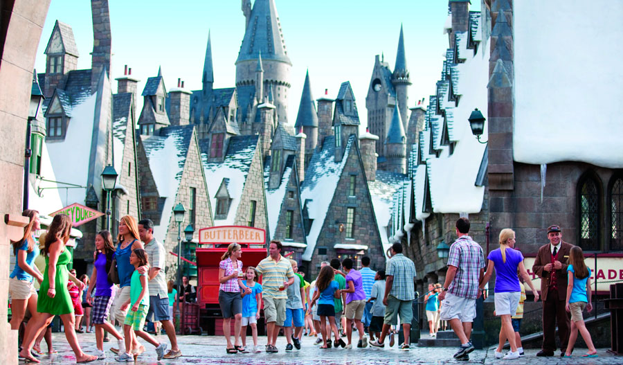 Pessoas caminham pela rua da área de Hogsmeade no parque temático de Harry Potter em Orlando, com construções bastante verticais que simulam textura de pedra e neve no topo. Ao fundo, vê-se o Castelo de Hogwarts, com suas torres icônicas.