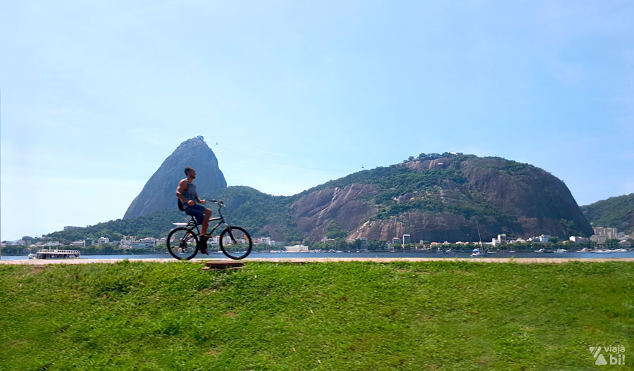 Homem andando de bicicleta em ciclovia com laterais gramadas. Ao fundo, os morros formam a paisagem icônica do Rio.