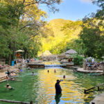 Pessoas relaxam em piscina aquecida com água esverdeada em meio à natureza. Ao fundo, árvores e montanhas, além de bares molhados