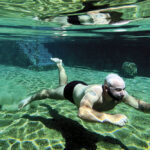 Foto subaquática com Rafael nadando em água cristalina esverdeada, com reflexo do sol no fundo da piscina